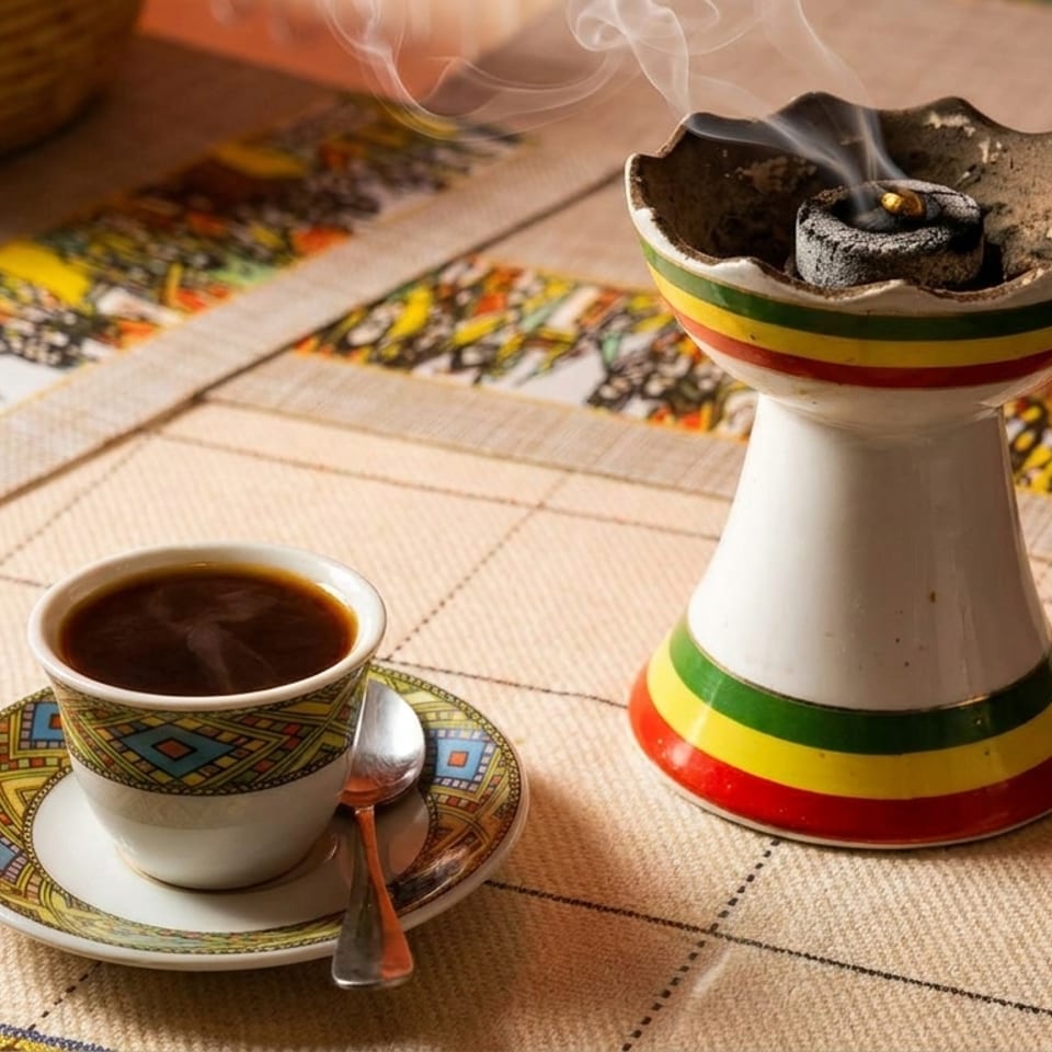 Steaming coffee in a patterned cup next to a smoking incense burner on a colorful tablecloth.