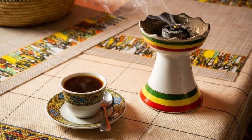 Steaming coffee in a patterned cup next to a smoking incense burner on a colorful tablecloth.